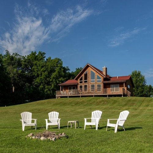 A wooden house on a hill with a deck, surrounded by green grass and four white chairs arranged around a fire pit.