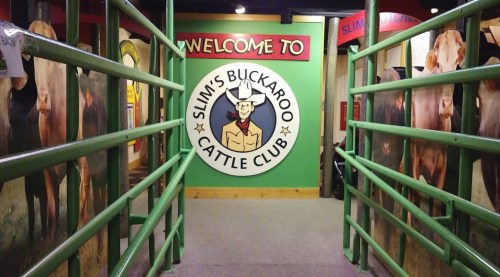 Entrance to Slim's Buckaroo Cattle Club, featuring a colorful sign and green railings with cow images on the walls.