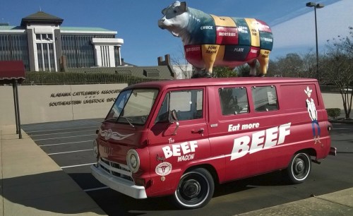 A vintage red food truck with a cow statue on top, promoting beef, parked outside a building.
