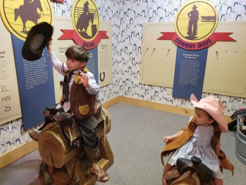 Two children dressed as cowboys sit on toy saddles, one waving a hat, with cowboy-themed displays in the background.