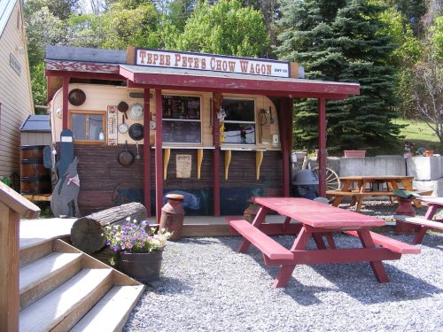 A rustic food stand with a red roof, picnic tables, and a menu board, surrounded by greenery and gravel.