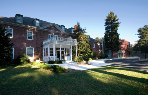A brick building with a porch, surrounded by greenery and trees, on a sunny day.