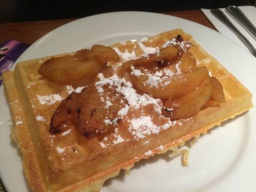 A golden waffle topped with caramelized apples and dusted with powdered sugar, served on a white plate.