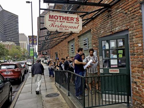 A line of people waiting outside Mother's Restaurant, a brick building with a sign, in an urban setting.