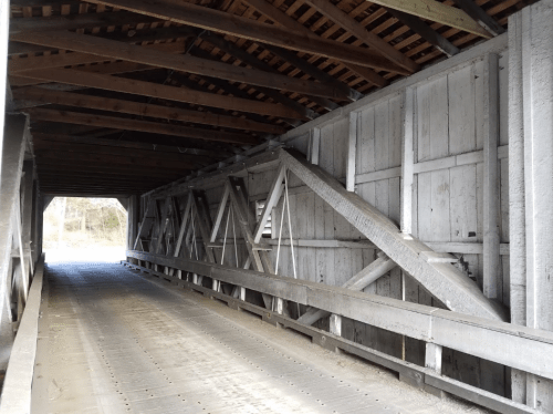 Interior view of a wooden covered bridge, showcasing beams and a wooden floor, with light at the far end.