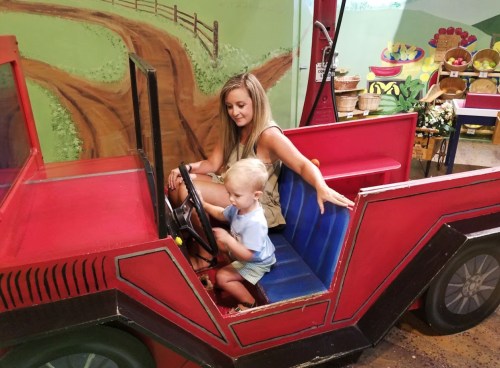 A woman and a young child sit in a colorful toy car, with the child pretending to drive.
