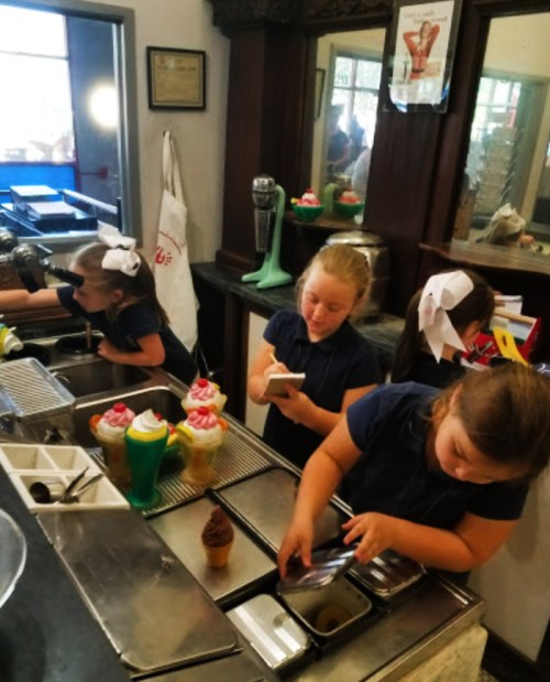 Three girls in blue shirts work at an ice cream shop, preparing treats and using a phone, with colorful desserts displayed.