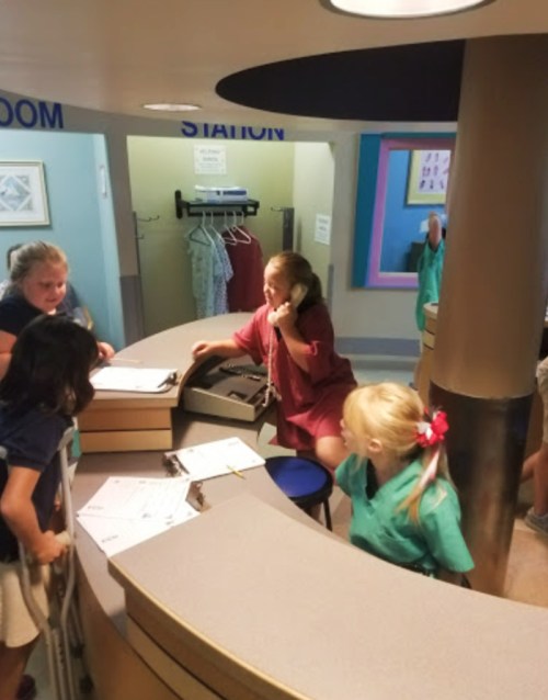 A group of children interact at a reception desk in a colorful room, with one child on the phone and others observing.