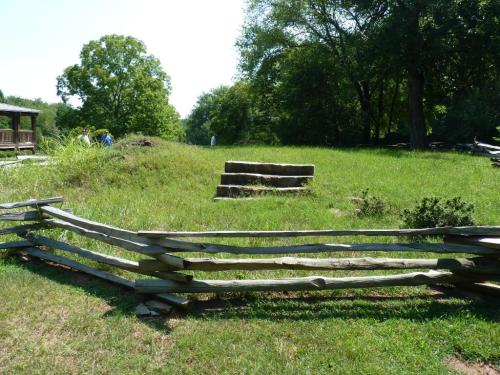 A grassy area with wooden steps and a split-rail fence, surrounded by trees and people in the background.