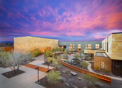 Modern building complex with warm-toned architecture, surrounded by desert landscaping under a vibrant sunset sky.