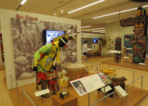 A colorful display featuring a performer playing a big drum in a museum, surrounded by Indigenous artifacts and information.
