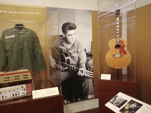 Elvis Presley’s military jacket, guitar, and a photo of him playing, displayed in a museum setting.
