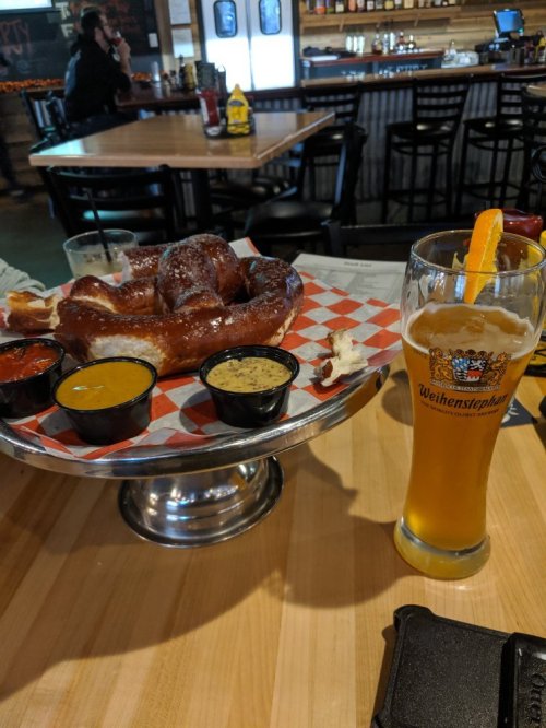 A large pretzel with dipping sauces on a plate, next to a tall glass of beer garnished with an orange slice.