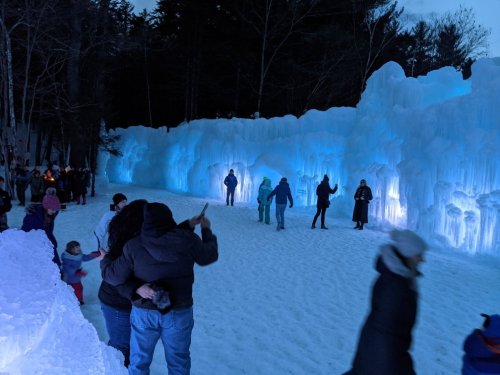 A winter scene with people exploring illuminated ice formations at dusk, surrounded by snow and trees.