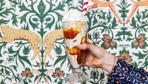 A hand holds a tall glass of ice cream topped with whipped cream and caramel, against a decorative patterned background.