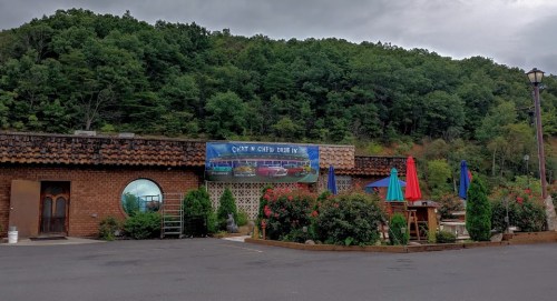 A rustic building with a mural, surrounded by greenery and colorful umbrellas, set against a hilly backdrop.