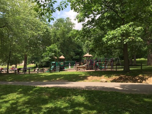 A playground surrounded by trees, featuring play structures and swings, with a path and grassy area in the foreground.