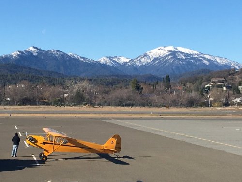 A bright orange airplane on a runway with snow-capped mountains in the background under a clear blue sky.