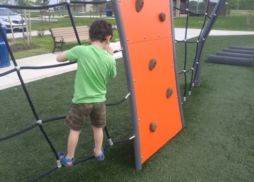 A child in a green shirt climbs a play structure with an orange wall at a park.
