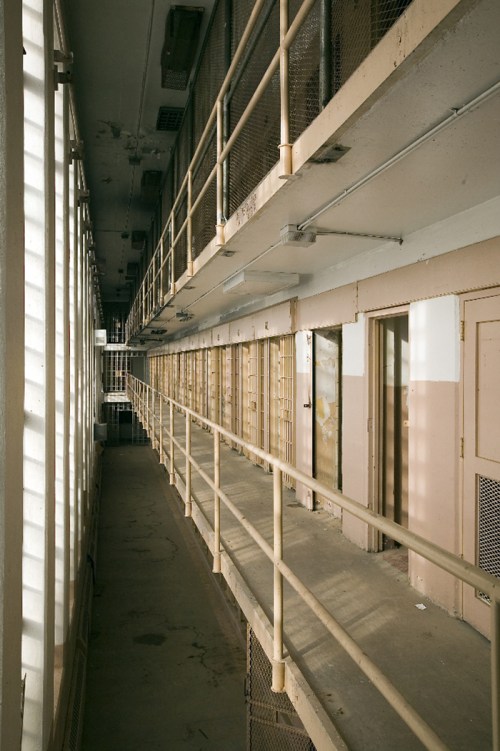 A long, empty prison corridor with metal railings and closed cell doors on either side, illuminated by natural light.