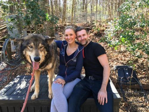 A woman and a man sit on a platform with a large wolf beside them, surrounded by trees and greenery.