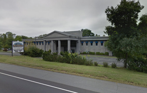 A large, gray building with columns and blue windows, surrounded by greenery, viewed from the roadside.