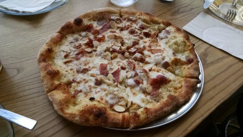 A freshly baked pizza topped with cheese, sliced meats, and almonds on a wooden table.