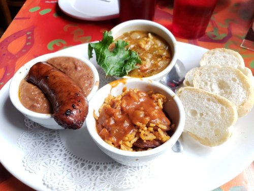 A plate featuring sausage, rice with sauce, beans, a bowl of soup, and slices of bread, garnished with lettuce.