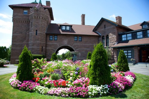 A grand stone mansion with a turret, surrounded by a vibrant flower garden in full bloom under a clear blue sky.