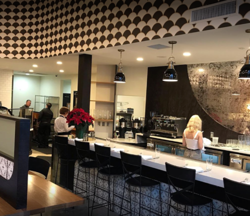 A modern restaurant interior featuring a bar, seating, and staff preparing food in the background.