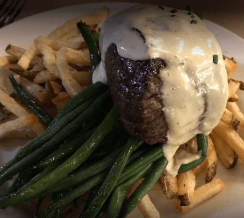 A plate of steak topped with creamy sauce, served on a bed of green beans and French fries.