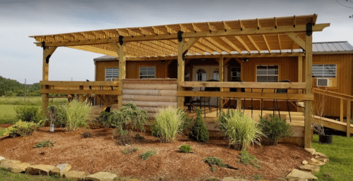 A wooden deck with a pergola, surrounded by landscaped plants, in front of a rustic cabin.