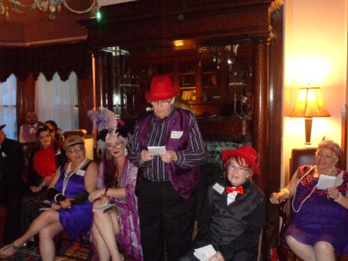 A group of people in vintage attire, sitting in a cozy room with elegant decor, engaged in conversation.