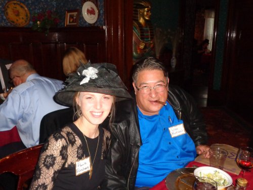 A woman in a black hat and a man with a cigar smile at a dinner table, surrounded by a festive atmosphere.