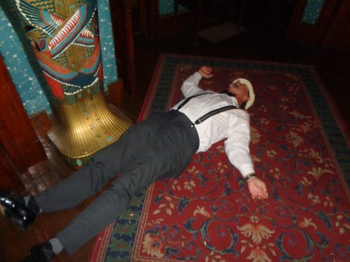 A man in formal attire lies on a patterned rug, appearing to be unconscious or resting, in a dimly lit room.