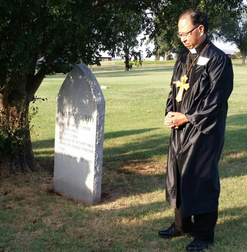 A person in a black robe stands solemnly by a gravestone in a grassy area, with a tree in the background.