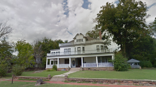 A large, two-story white house with a wraparound porch, surrounded by trees and a grassy lawn under a cloudy sky.