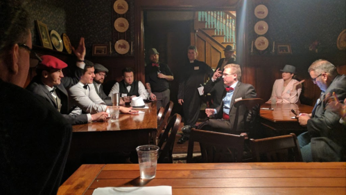 A group of people in formal attire gathered around a wooden table in a dimly lit room, engaged in conversation.