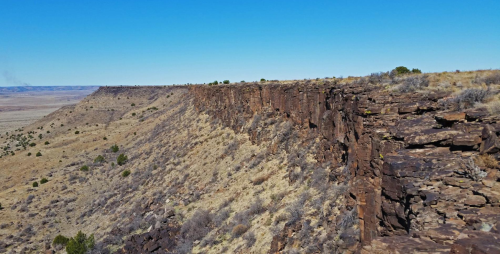 A rocky cliff edge overlooking a vast, arid landscape under a clear blue sky. Sparse vegetation is visible.