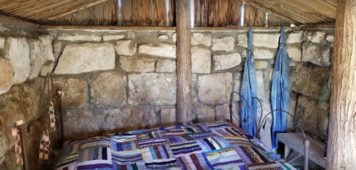Interior of a rustic room with stone walls, a quilted bed, and hanging denim garments.