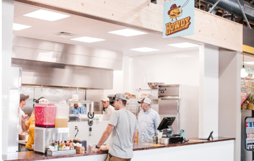 A busy burger restaurant with staff preparing food and a customer at the counter, featuring a colorful drink station.