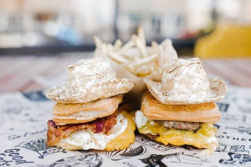 Two sandwiches wearing tiny straw hats, served with a side of fries on a patterned paper background.