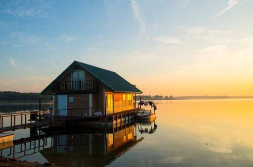 A serene lakeside house at sunrise, reflecting on calm waters with a boat docked nearby.