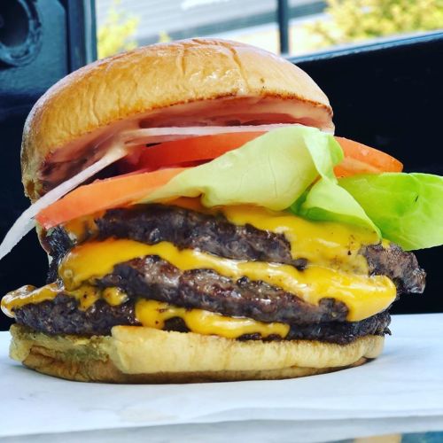 A close-up of a stacked cheeseburger with three beef patties, melted cheese, lettuce, tomato, and onion on a toasted bun.