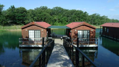 Two wooden cabins on stilts by a calm lake, connected by a wooden walkway, surrounded by lush greenery.