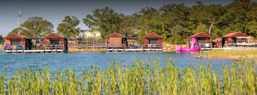 A serene lakeside scene featuring wooden cabins, a pink float, and lush greenery along the water's edge.