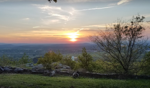 A scenic sunset over rolling hills, with trees and rocks in the foreground, casting a warm glow across the landscape.
