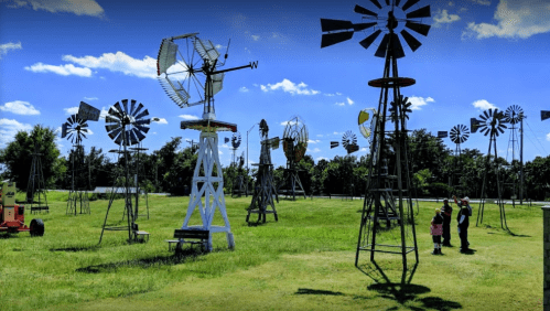 A field with various vintage windmills under a bright blue sky, with two people observing the scene.