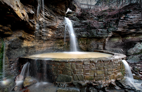 A circular stone pool with water flowing from a rocky cliff, surrounded by lush greenery and a serene atmosphere.