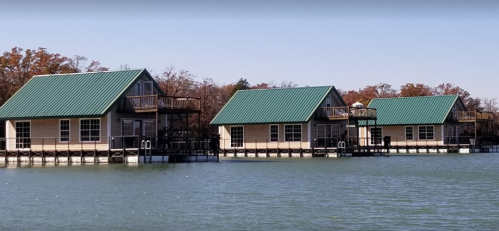 Three lakeside cabins with green roofs on stilts, surrounded by calm water and autumn trees in the background.